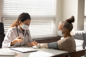 Caring female nurse in medical facial mask measure check blood pressure of mature woman patient....