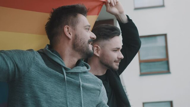 Proud young gay male couple waving rainbow flag from the balcony. High quality 4k footage