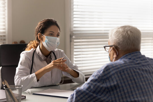 Young female GP in medical facemask consult mature male patient during corona virus quarantine in hospital. Woman doctor in facial mask talk on consultation with elderly client. Healthcare concept.