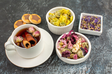 sidewise view of a cup of herbal tea with cookies and three bowls of dried flowers on grey ground