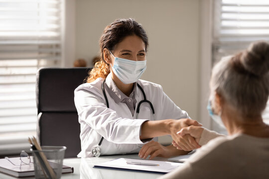 Smiling Female GP Or Physician In Medical Facemask Handshake Mature Patient At Hospital Meeting. Happy Woman Doctor In Facial Mask Shake Hand Close Health Insurance Deal With Elderly Client In Clinic.