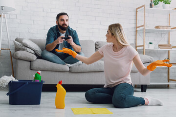Disgruntled woman in rubber gloves spreads her arms to sides and looks at husband playing at...