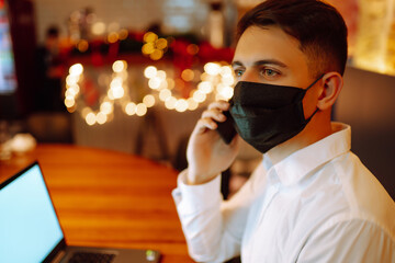 Young man wearing medical face mask working on laptop computer  and speaks on the phone at home during winter holiday. The concept of preventing the spread of the epidemic coronavirus. Remote work. 