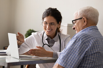 Happy female doctor and senior male patient look at computer screen consider good results after...