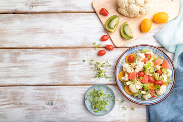Vegetarian salad of cauliflower cabbage, kiwi, tomatoes, microgreen sprouts on gray wooden background. Top view, copy space.