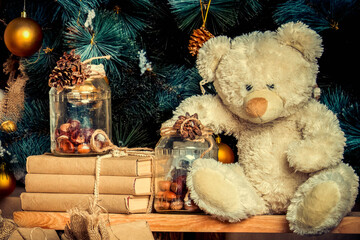 Close up beige teddy bear under christmas tree. Christmas decoration with vintage toys, golden balls, books in craft wrapper, glass jar with pine cones. vintage toning. Present for child on New Year