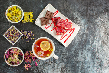 top view of a cup of herbal tea with bowls of dry flowers and a plate of chocolate on grey ground