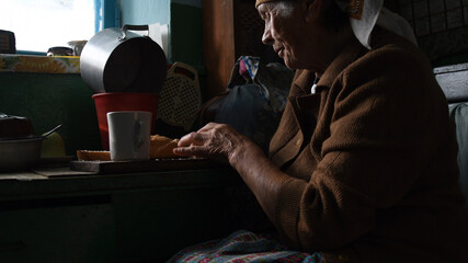 Old wrinkled woman tilts her head and takes bread by hand. Suffering loneliness at senior age. Portrait of rural elderly woman in contour sunlight. Concept of poverty and geriatric health problem