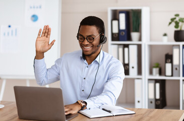 Happy black consultant with headset waving at laptop screen