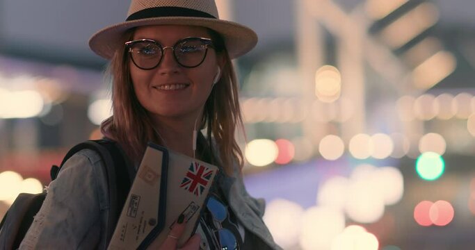 Happy hipster young woman in stylish glasses and hat wear headphones and listening music with blurred night city and street lights on background. Smiling model dancing alone. People and style concept