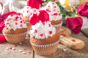 Cupcake with Heart for Valentines. Valentine Day Sweet Dessert, vanilla cupcakes with whipped vanilla cream and red sugar hearts decor for Valentine's Day, wooden background with rose flowers bouquet