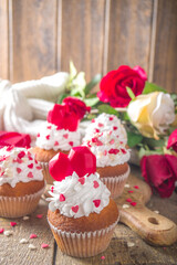 Cupcake with Heart for Valentines. Valentine Day Sweet Dessert, vanilla cupcakes with whipped vanilla cream and red sugar hearts decor for Valentine's Day, wooden background with rose flowers bouquet