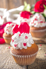 Cupcake with Heart for Valentines. Valentine Day Sweet Dessert, vanilla cupcakes with whipped vanilla cream and red sugar hearts decor for Valentine's Day, wooden background with rose flowers bouquet