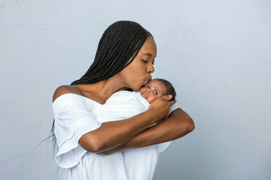 A beautiful young African-American woman with braids is kissing her newborn son and looking at him with love on a white gray background and giving him a kiss