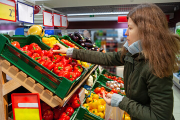 Woman holding red pepper, examining it with care, while grocery shopping in supermarket, standing in front of boxes filled with fresh fruits and vegetables. Woman buying bell peppers at grocery store.