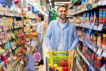 Young cheerful man shopping in supermarket with trolley cart