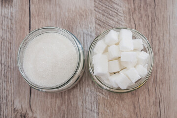 Two different types of sugar (white sand and sugar cubes) in jars, wood background.