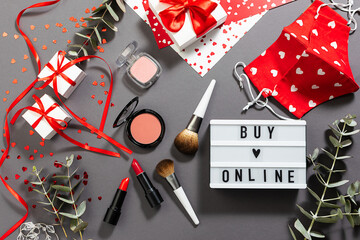 Creative St. Valentine’s Day composition with text Buy Online on the lightbox, set of decorative cosmetic, protective masks with heart-shaped print and shining decorations on trendy gray background.