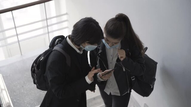 Top angle view of two high school female students in Covid-19 face masks surfing Internet on smartphone. Carefree Caucasian girls friends standing on stairs talking on coronavirus pandemic.