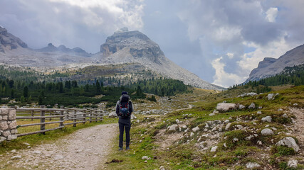 Fototapeta na wymiar A man with a big hiking backpack hiking along a gravelled road in Italian Dolomites. There is a wooden fence along the road. Sharp and stony mountain chains in front. Few boulders on the green meadow.