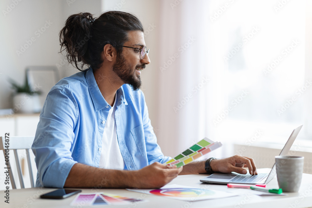 Wall mural young eastern interior designer holding color swatch samples and working on laptop