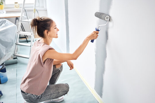 Smiling woman painting with paint roller on wall while crouching at home