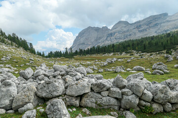 A stony wall made of bigger lose boulders on a lush green meadow in Italian Dolomites. There are sharp and stony mountains in the back. The mountains are surrounded by clouds. Remote and desolate area