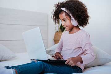 Cute girl using laptop wearing headphone listening music while sitting in bedroom at home