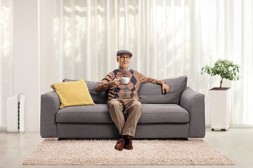 Elderly man seated on a sofa holding a cup and smiling