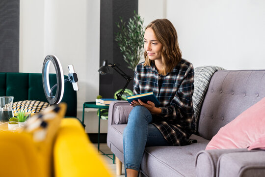 Smiling woman holding book while live streaming through mobile phone attached to ring tripod at home