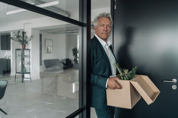 Businessman holding box of botany plant while leaning on glass wall at office