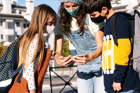 Mother And Kids Wearing Protective Face Mask Using Smart Phone Standing In Public Park On Sunny Day