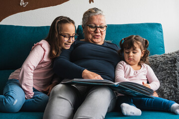 A grandmother with her two granddaughters, reading a book to them on the sofa at home.