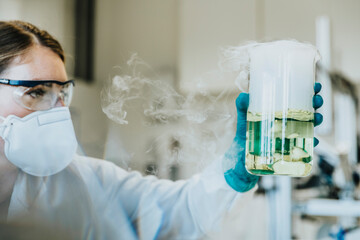 Laboratory assistant wearing protective face mask and eyeglasses holding liquid beaker while standing at laboratory