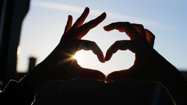 Hands forming a heart in front of a window during romantic sunset. Symbol of love, empathy, and altruism while staying at home. Concept of long-distance relationship and Valentine's day in quarantine.