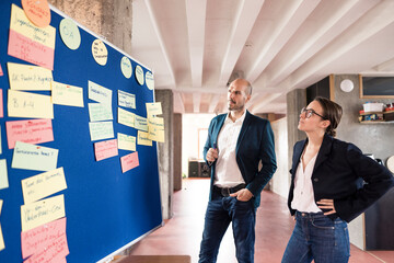 Businessman and woman reading note putted on bulletin board while standing at office