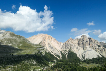 Fototapeta premium A panoramic view on the valley in Italian Dolomites. Dense forest overgrows the mountains slopes. Lush green plants. High mountain chains in the back. Spring in the mountains. Sunny day. Serenity