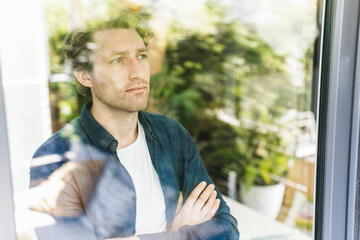 Mid adult man with arms crossed looking through window while standing at home