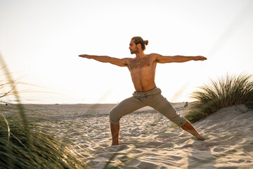 Shirtless man practicing warrior 2 position yoga at beach against clear sky during sunset