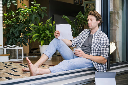 Mid adult man with coffee cup holding paper while sitting at home