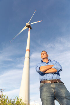 Confident mature male engineer standing with arms crossed against wind turbines on field