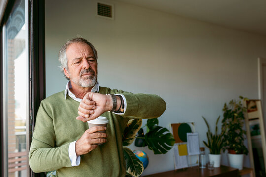 Man with coffee cup checking time while standing by window at home