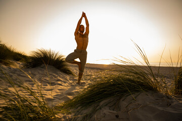 Shirtless man practicing tree pose amidst plants at beach against clear sky during sunset