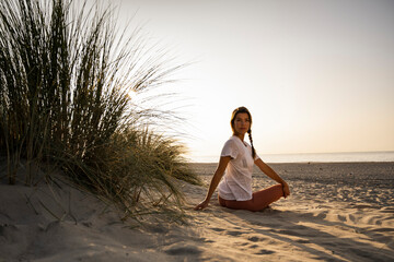 Beautiful young woman practicing yoga while sitting by plant at beach against clear sky during sunset