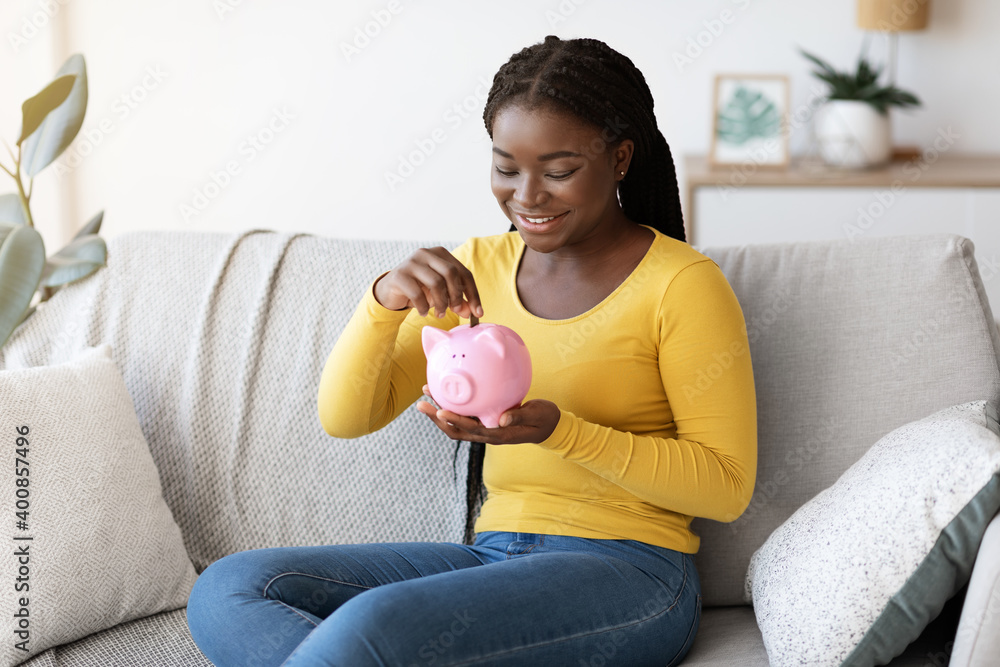 Wall mural Black woman putting coin in piggy bank, sitting on couch at home