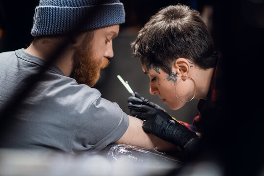 A tattoo artist with short hair and piercings is preparing to get a tattoo on a man's arm in a tattoo parlor.