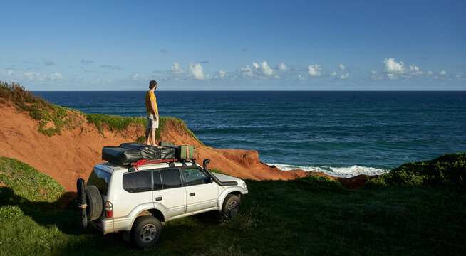 Male tourist standing on 4x4 roof while looking at sea against blue sky