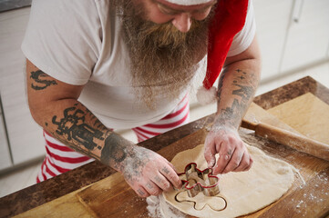 Top view of a man in Santa Claus hat and tattooed arms cutting a shape out of dough, baking a gingerbread man in the kitchen