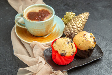 Front close view of a cup of frothy coffee on towel with accessory and small cupcakes on dark background