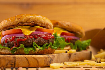 Appetizing burgers with beef, tomato, lettuce, red onion, cheese, mustard and ketchup on wooden chopped board with french fries.
